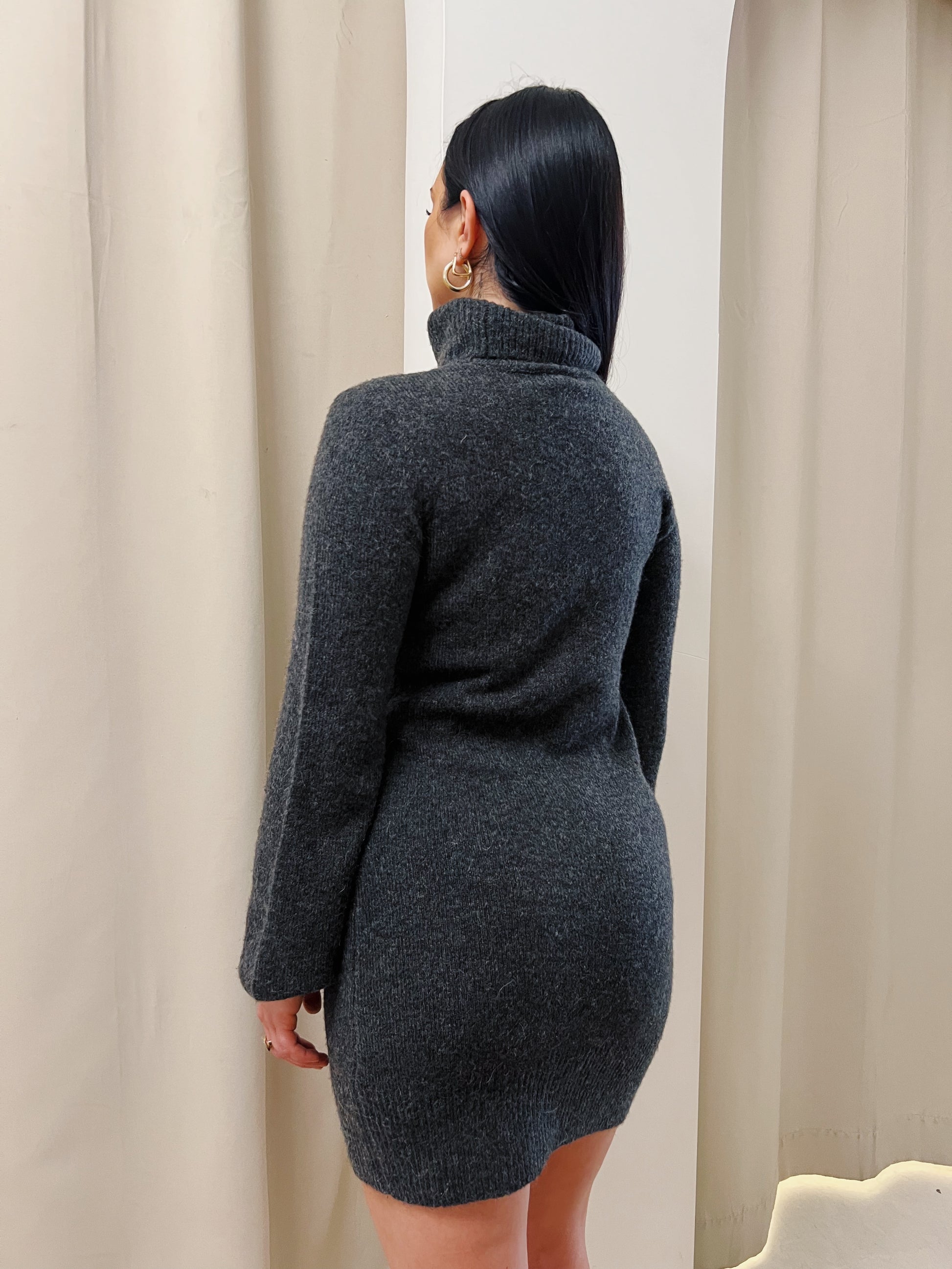 Woman is standing with her back facing the camera wearing a dark grey turtleneck dress